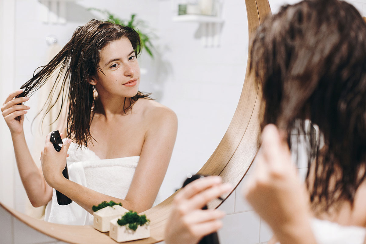 Молодая счастливая женщина в белом полотенце наносит маску-кондиционер на волосы в ванной